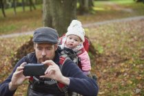 Mittlerer erwachsener Mann im Park trägt Schiebermütze mit Tochter auf dem Rücken in Tragetasche und macht Selfie mit Smartphone — Stockfoto