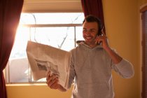 Молодой человек смотрит на газету и использует мобильный телефон в номере отеля, улыбаясь — стоковое фото