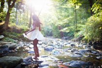 Glamorous young woman wearing white dress stepping over forest river rocks — Stock Photo