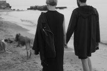 Jovem casal caminhando ao longo da praia de mãos dadas, dois cães correndo à frente, visão traseira — Fotografia de Stock