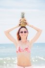 Портрет жінки середнього віку на пляжі, одягнена в сонцезахисні окуляри, тримає ананас на голові — стокове фото