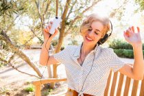 Mid adult woman sitting on bench, wearing headphones, holding smartphone, dancing — Stock Photo