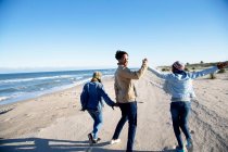 Tres amigos caminando por la playa, tomados de la mano, vista trasera - foto de stock
