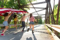 Grupo de amigos corriendo en el puente sosteniendo bandera americana - foto de stock