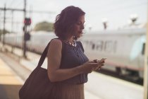 Mid adult woman waiting at train station, holding smartphone — Stock Photo