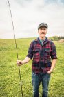 Jovem no campo vestindo camiseta e tampa plana segurando haste de pesca olhando para a câmera sorrindo — Fotografia de Stock