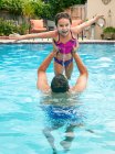 Padre en la piscina levantando a su hija, brazos abiertos mirando a la cámara sonriendo - foto de stock
