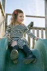Cute girl in striped dress sitting on playground slide — Stock Photo