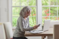 Старшая женщина использует смартфон и ноутбук дома — стоковое фото