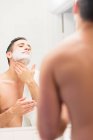 Середній дорослий чоловік, дивлячись у дзеркало, наносить піну для гоління на шию, вид ззаду — стокове фото