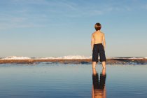 Vista posteriore del ragazzo e riflessione in piedi sulla spiaggia — Foto stock