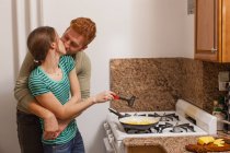 Молодий чоловік в кухонних обіймах навколо молодої жінки готує на холодильнику, цілує — стокове фото