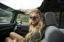 Jovem mulher com cabelo loiro longo varrido pelo vento na estrada em jipe — Fotografia de Stock