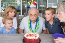 Senior man blowing out candles on birthday cake with family — Stock Photo