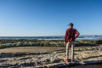 Молодой человек стоит на пляже и смотрит на океан, вид сзади — стоковое фото
