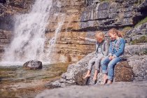 Hermano y hermana, sentados en la roca, relajados, junto a la cascada - foto de stock