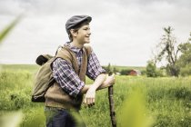 Side view of teen boy in field wearing flat cap leaning against branch looking away smiling — Stock Photo