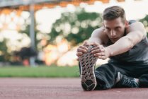 Young man on sports track, holding foot, stretching — Stock Photo