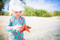 Girl on beach wearing swimwear and sunhat holding starfish looking down, St. Croix, US Virgin Islands — Stock Photo