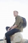 Молодой человек в рюкзаке сидит на скале и смотрит в сторону — стоковое фото