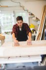 Jeune homme en atelier portant un tablier en utilisant une scie à table pour couper le bois — Photo de stock