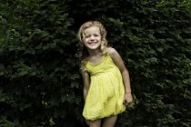 Portrait of smiling blond haired girl wearing yellow dress standing in front of garden hedge — Stock Photo