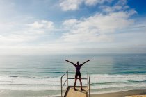 Jovem mulher de pé em olhar para fora ponto ao lado do mar, braços levantados, visão traseira — Fotografia de Stock