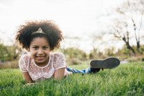 Vista a nivel de la superficie de la niña acostada en el frente en la hierba sonriendo, mirando a la cámara - foto de stock