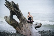 Mature woman practicing yoga lotus position on large driftwood tree trunk at beach — Stock Photo