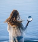 Rear view of young woman with long red hair standing in lake holding crystal ball — Stock Photo