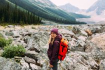 Vue latérale d'une femme adulte sur un paysage rocheux portant un sac à dos regardant une caméra souriante, lac Moraine, parc national Banff, Alberta Canada — Photo de stock
