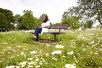 Зрелая женщина сидит на скамейке в парке, заполненном ромашками — стоковое фото