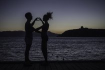 Casal fazendo forma de coração com as mãos por mar, silhueta — Fotografia de Stock