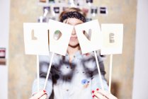 Young woman in front of photo wall hiding behind love sign — Stock Photo