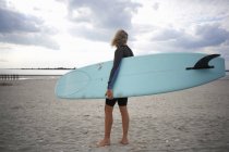Старшая женщина стоит на пляже, держа доску для серфинга, вид сзади — стоковое фото