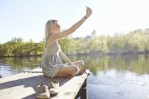 Young woman sitting on jetty photographing herself — Stock Photo