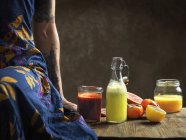 Cropped view of  woman wearing dress sitting on table with raw juices in glass bottle and jars — Stock Photo