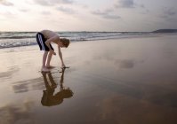 Menino curvando-se sobre tocar areia na praia — Fotografia de Stock