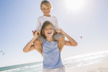 Большой брат на пляже таскает мальчика на плечах улыбаясь — стоковое фото