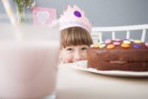 Girl in crown hiding behind birthday cake on table — Stock Photo
