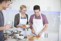 Mittlerer erwachsener Mann hackt Möhren, während Freunde in der Küche zuschauen — Stockfoto
