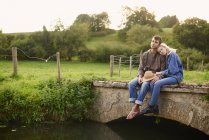 Romantique jeune couple assis sur passerelle de rivière — Photo de stock