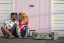 Young couple sitting by beach hut with suitcase — Stock Photo