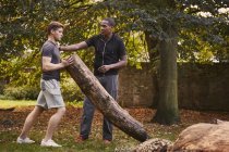 Young man with personal trainer lifting tree trunk in park — Stock Photo