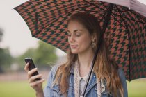 Молодая женщина с зонтиком смс на смартфоне в парке — стоковое фото