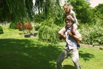 Hombre maduro corriendo con hija sobre sus hombros en el jardín - foto de stock