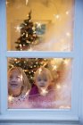 Hermanas mirando por la ventana con decoraciones navideñas - foto de stock