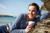 Retrato de jovem na costa com café e fones de ouvido — Fotografia de Stock