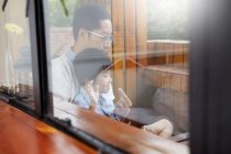 Pai chinês e filho jovem tomando café da manhã na varanda ao sol juntos, baleado pela janela — Fotografia de Stock