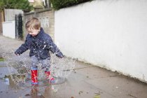 Male toddler in red rubber boots splashing in sidewalk puddle — Stock Photo
